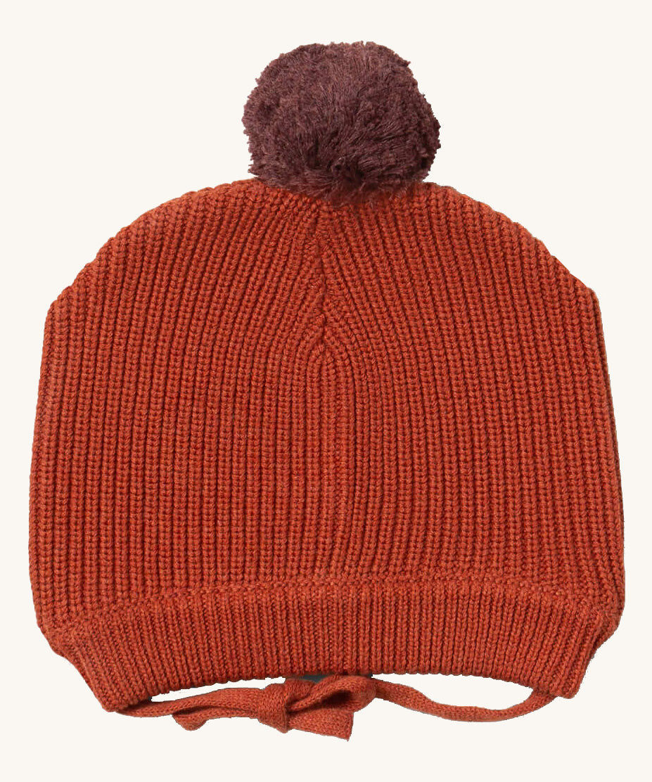Little Green Radicals ginger knitted beanie hat available at Babipur. A dark orange knitted GOTS organic cotton beanie style hat with a purple pom pom on top. Features matching dark orange ties for a secure fit.