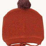 Little Green Radicals ginger knitted beanie hat available at Babipur. A dark orange knitted GOTS organic cotton beanie style hat with a purple pom pom on top. Features matching dark orange ties for a secure fit.