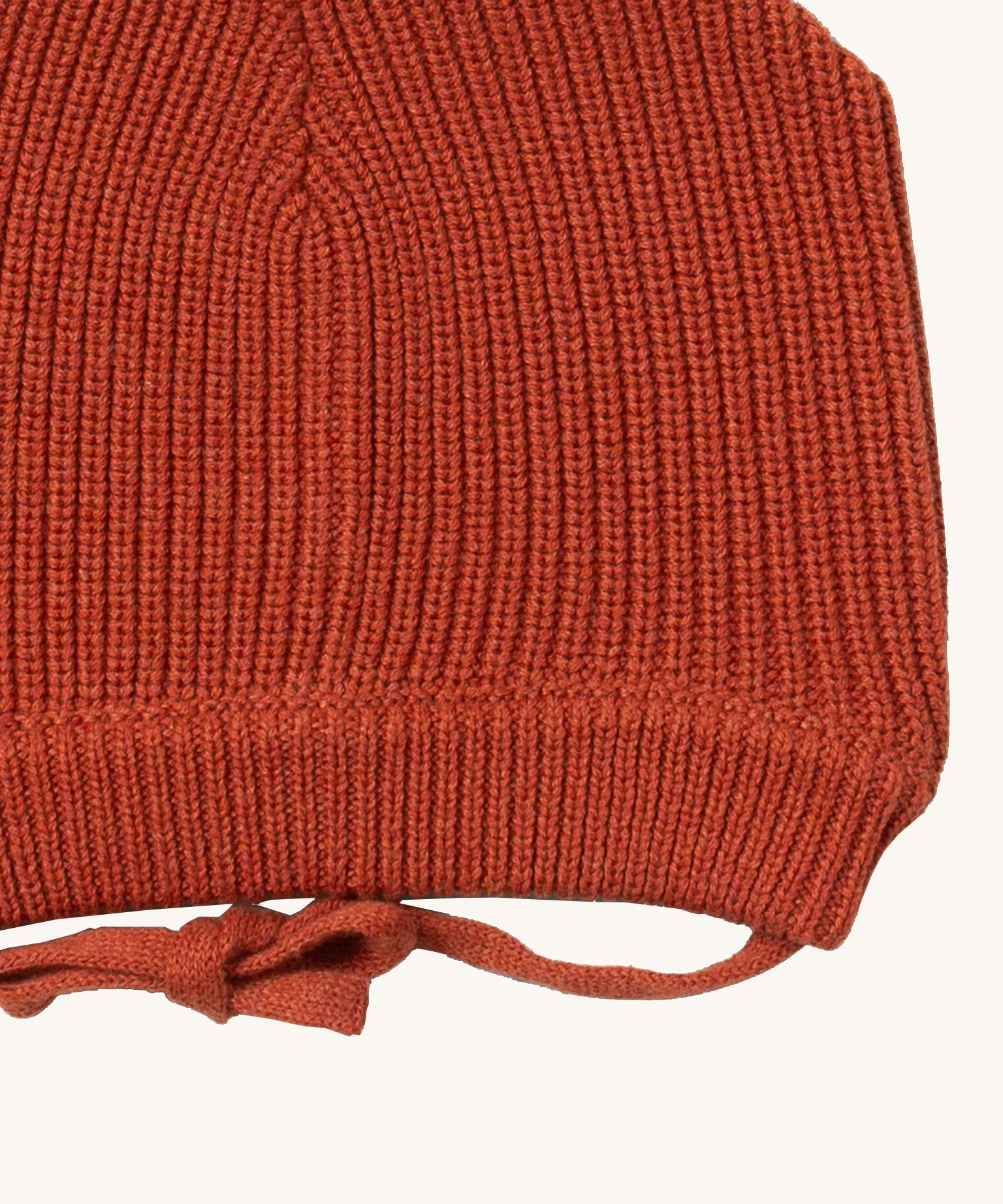 A close up of the Little Green Radicals ginger knitted beanie hat showing the closed dark orange tie detail on organic cotton fabric