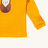 A close up of the Little Green Radicals gold brown owl applique long sleeve top showing the cute brown owl applique and sleeve detail on dark yellow organic cotton fabric