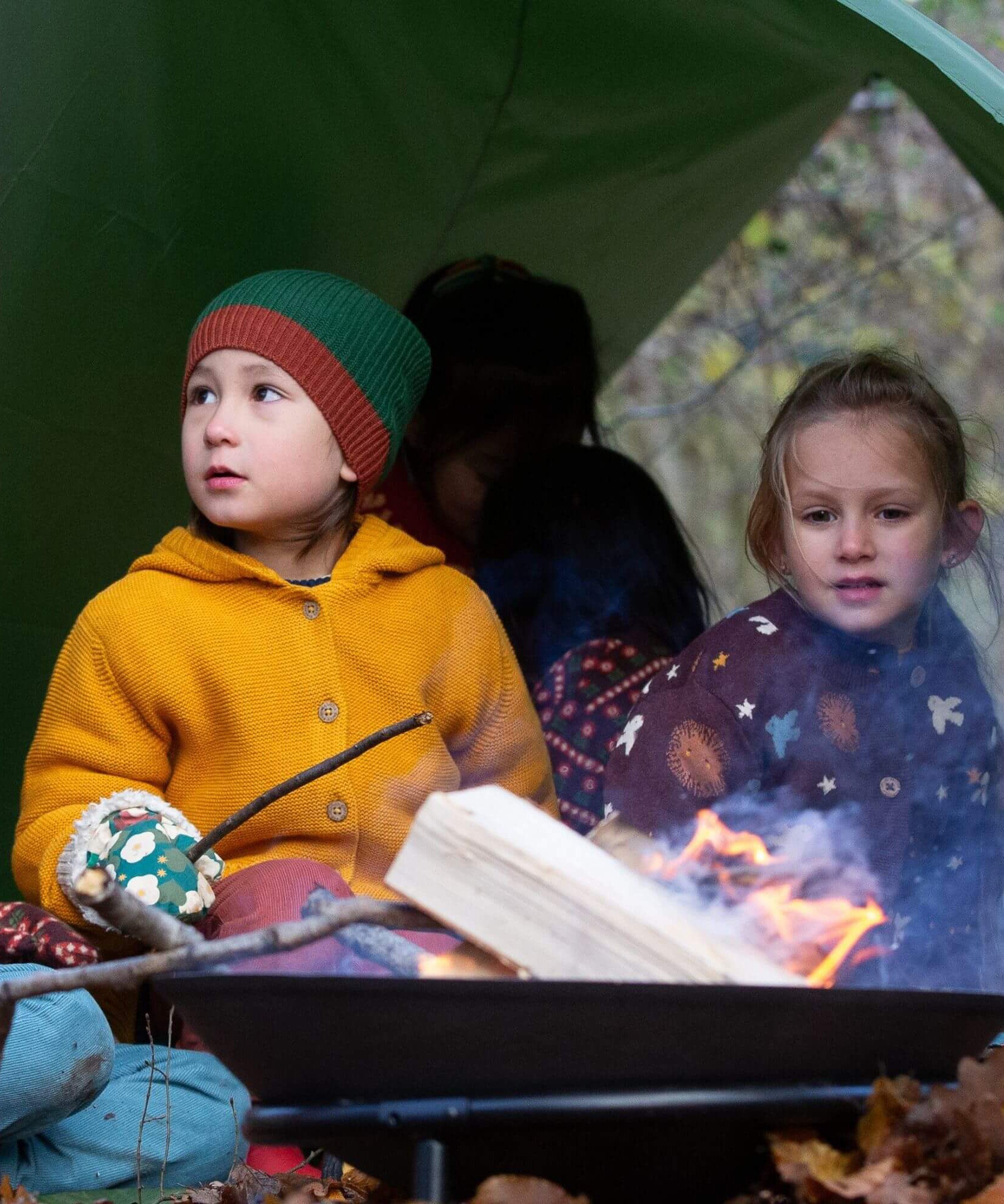 A child sitting under a shelter in woodland wearing the Little Green Radicals gold snuggly hooded knitted cardigan. A thick knitted cardigan with a generous hood for warmth and button fasteners  down the middle on yellow organic cotton fabric. 