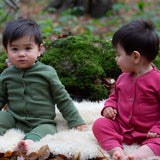 2 children wearing the holly berry pink and green chive waffle playsuit outside in woodland. A long sleeved, footless all in one suit made from GOTS organic cotton fabric with button fasteners and a small pouch chest pocket
