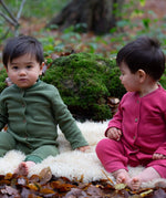 2 children wearing the holly berry pink and green chive waffle playsuit outside in woodland. A long sleeved, footless all in one suit made from GOTS organic cotton fabric with button fasteners and a small pouch chest pocket