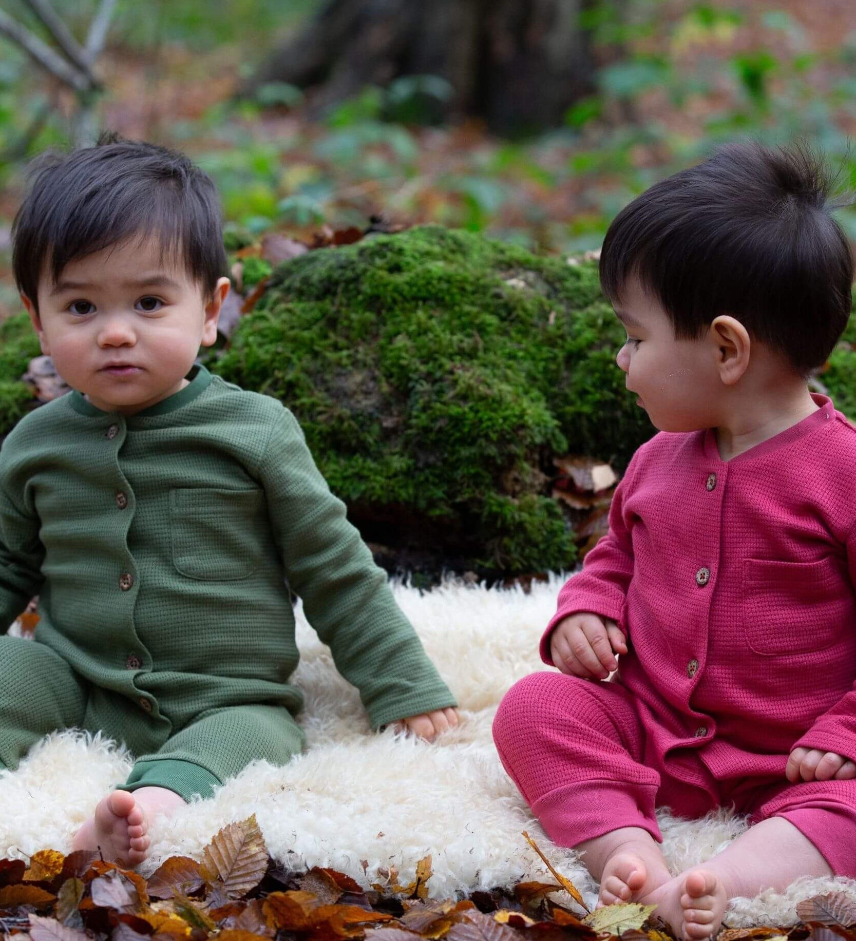 2 children wearing the holly berry pink and green chive waffle playsuit outside in woodland. A long sleeved, footless all in one suit made from GOTS organic cotton fabric with button fasteners and a small pouch chest pocket