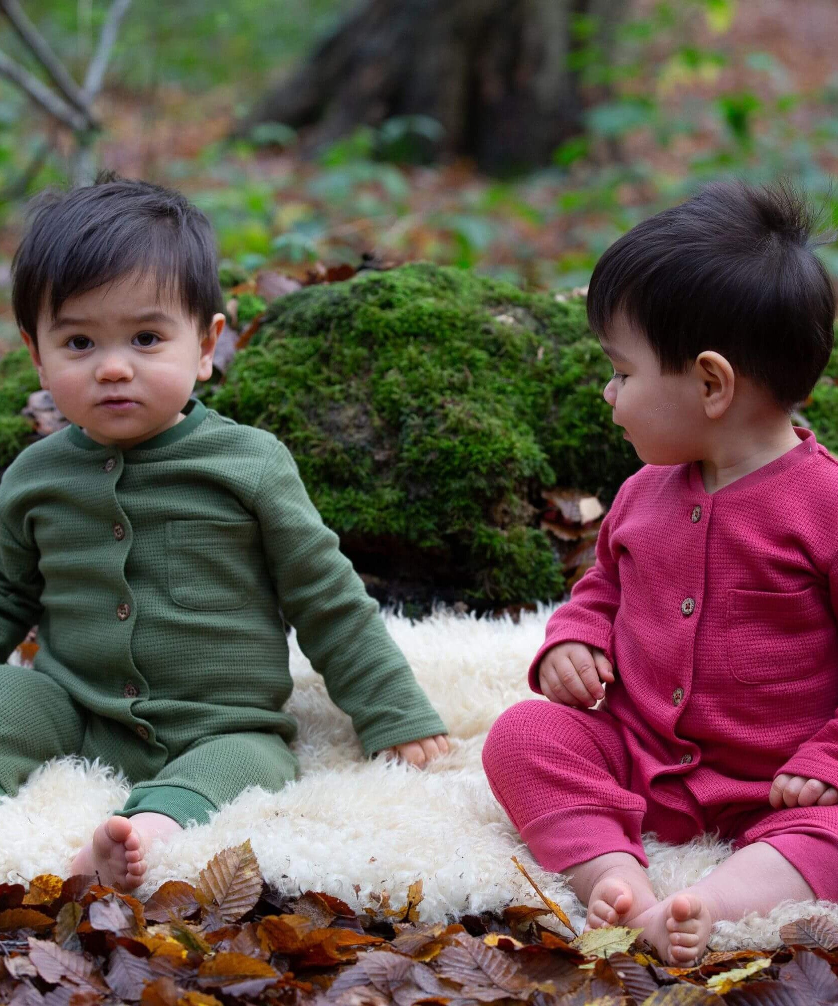 2 children wearing the holly berry pink and green chive waffle playsuit outside in woodland. A long sleeved, footless all in one suit made from GOTS organic cotton fabric with button fasteners and a small pouch chest pocket