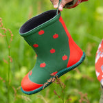 A Little Green Radicals kids green wellington boots held by a child's hand.  Showing the red apples prints all over the natural rubber waterproof boot.  Features a red toe cap, gripped sole and pull-on tab for easy fitting