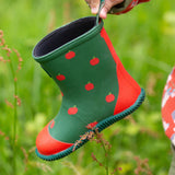 A Little Green Radicals kids green wellington boots held by a child's hand.  Showing the red apples prints all over the natural rubber waterproof boot.  Features a red toe cap, gripped sole and pull-on tab for easy fitting
