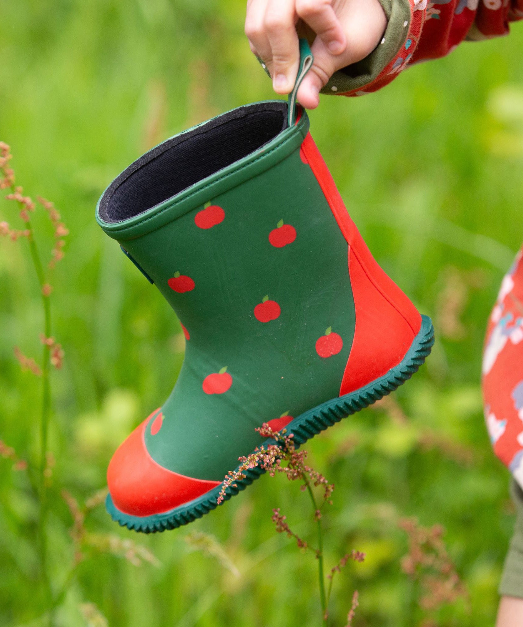 A Little Green Radicals kids green wellington boots held by a child's hand.  Showing the red apples prints all over the natural rubber waterproof boot.  Features a red toe cap, gripped sole and pull-on tab for easy fitting