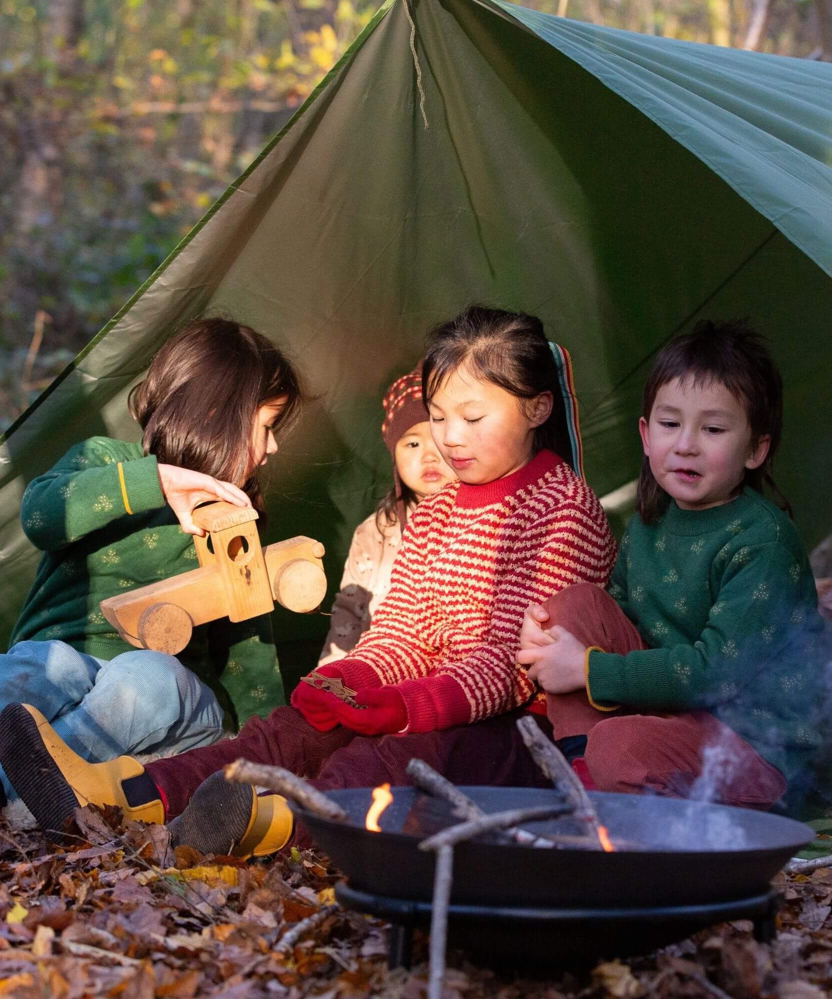 A group of kids sitting outside under a shelter including a child wearing the Little Green Radicals snuggly knitted jumper in juniper stripe. Available at Babipur this thick warm knitted jumper  comes in a yellow and pale red stripe design. Features a extendable arm cuff for size adjusting. 