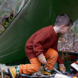 A child wearing the Little Green Radicals ginger aran knitted jumper in woodland under a shelter. A thick cable knit style jumper in dark orange GOTS organic cotton fabric with striped cuffs.