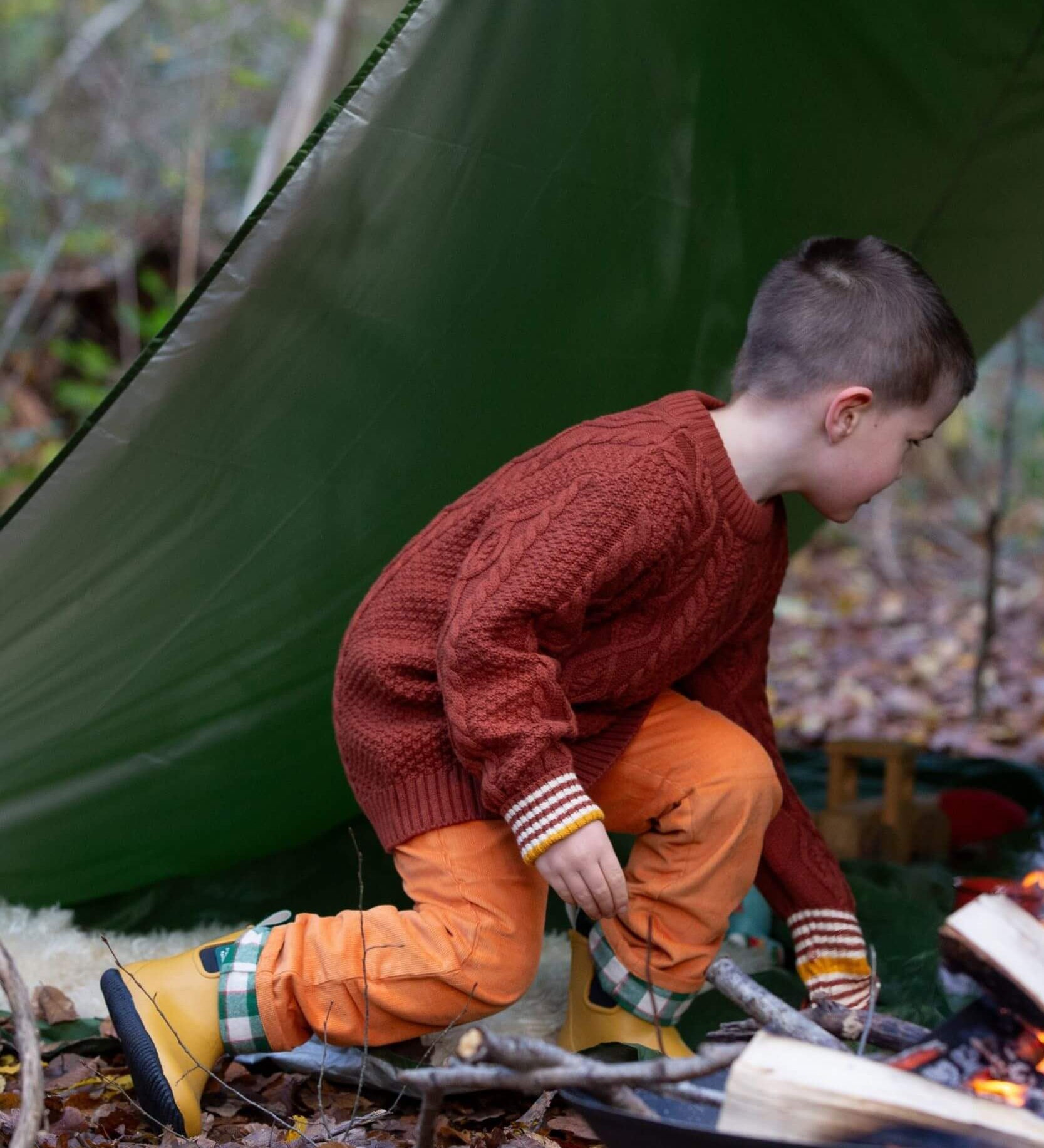 A child wearing the Little Green Radicals ginger aran knitted jumper in woodland under a shelter. A thick cable knit style jumper in dark orange GOTS organic cotton fabric with striped cuffs.