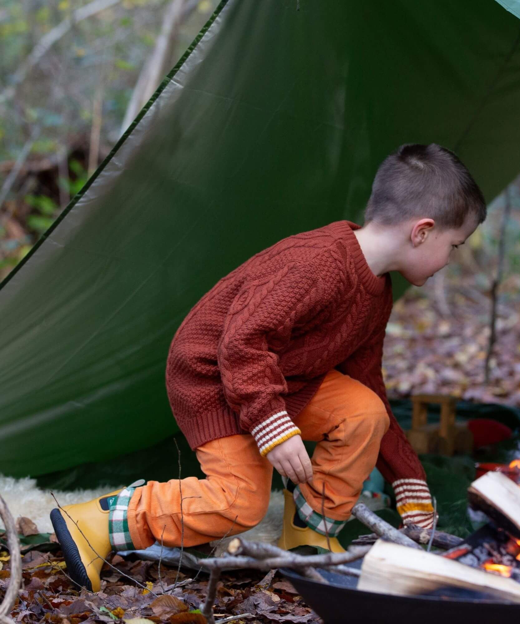 A child wearing the Little Green Radicals ginger aran knitted jumper in woodland under a shelter. A thick cable knit style jumper in dark orange GOTS organic cotton fabric with striped cuffs.