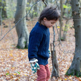 A child standing in woodland wearing the Little Green Radicals navy aran knitted jumper. A thick cable knit style jumper in dark navy GOTS organic cotton fabric with striped cuffs and a thick collar for warmth