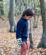 A child standing in woodland wearing the Little Green Radicals navy aran knitted jumper. A thick cable knit style jumper in dark navy GOTS organic cotton fabric with striped cuffs and a thick collar for warmth
