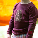 A close up of a child standing outside and wearing the dark maroon Little Green Radicals teddy bear applique long sleeve t shirt showing the bear and apple applique detail.