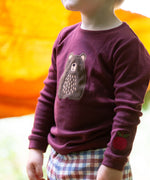 A close up of a child standing outside and wearing the dark maroon Little Green Radicals teddy bear applique long sleeve t shirt showing the bear and apple applique detail.