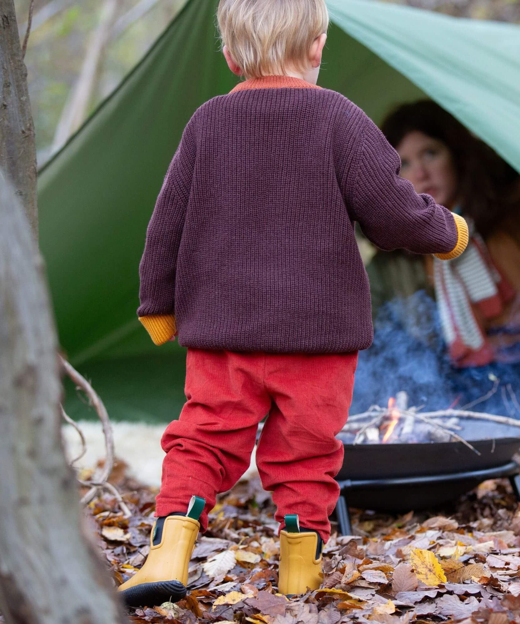A child walking through leaves in a woodland and wearing the Little Green Radicals comfy corduroy trousers in ginger with the navy sheep knitted jumper.  A corduroy style trouser that's loose fitted in dark orange organic cotton fabric