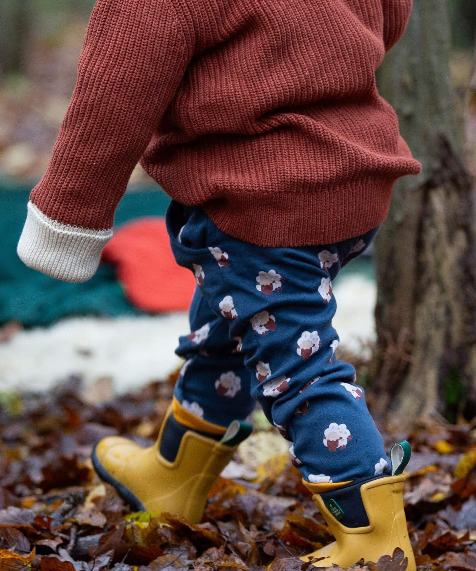 A child walking though leaves in a woodland wearing the Little Green Radicals comfy joggers in sleeping sheep. A navy jogger with cute prints of sheep on soft GOTS organic cotton fabric