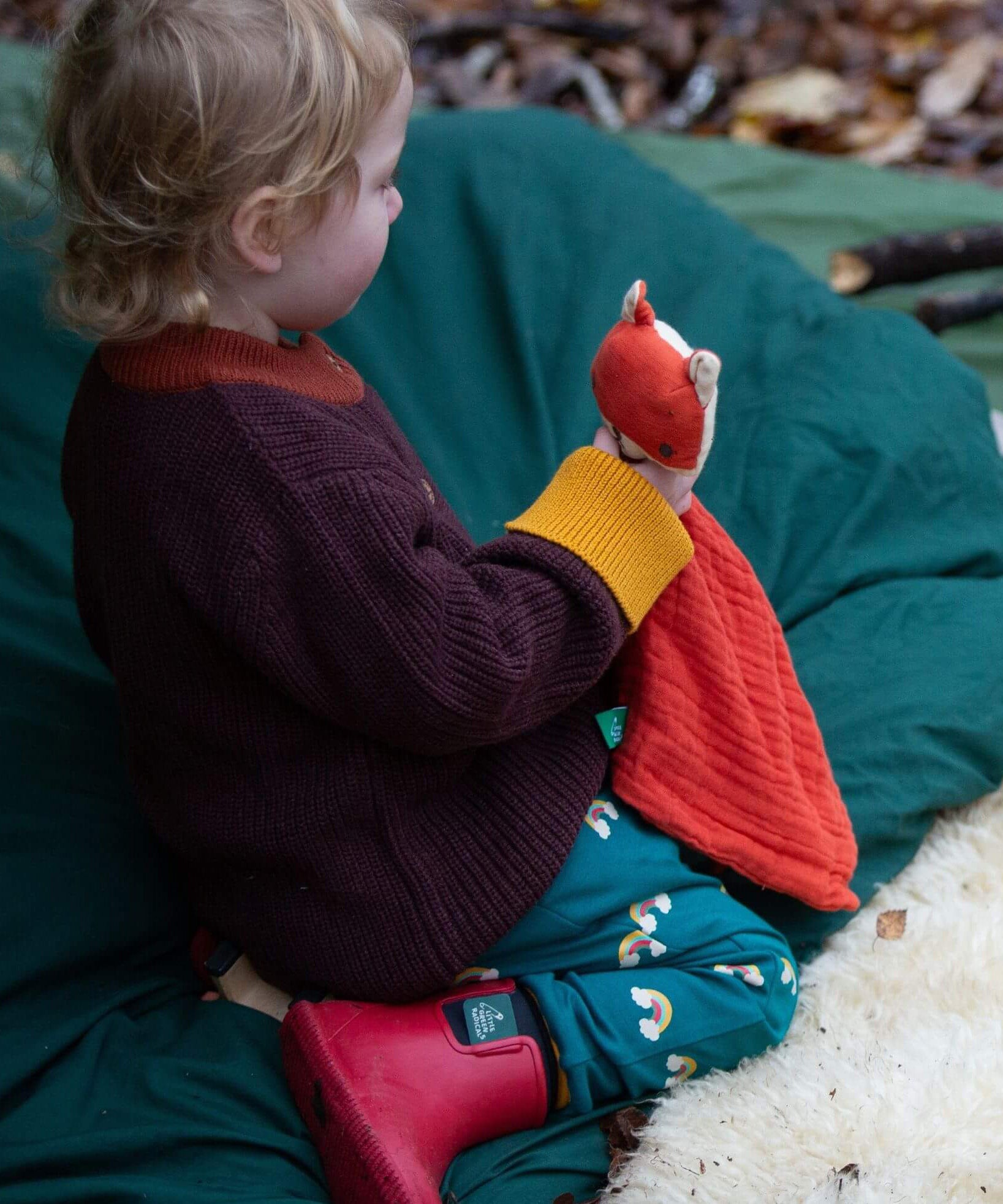 A child sitting down and wearing the Little Green Radicals turquoise rainbow joggers. A soft GOTS organic cotton jogger with cute prints of rainbows.