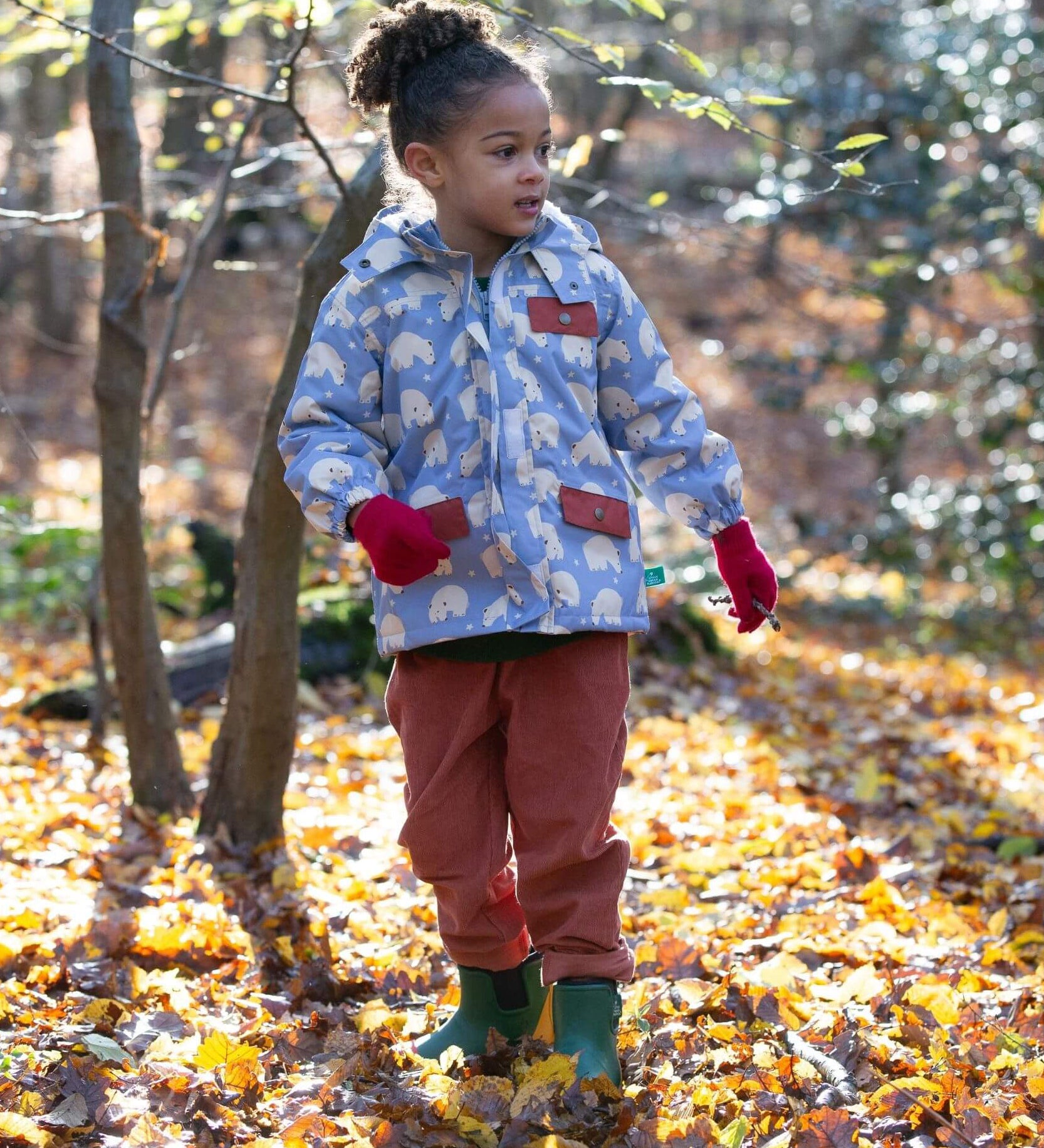 A child walking through leaves in a woodland playing with sticks and wearing the Little Green Radicals comfy corduroy trousers in ginger.  A corduroy loose fitted trousers in dark orange organic cotton fabric