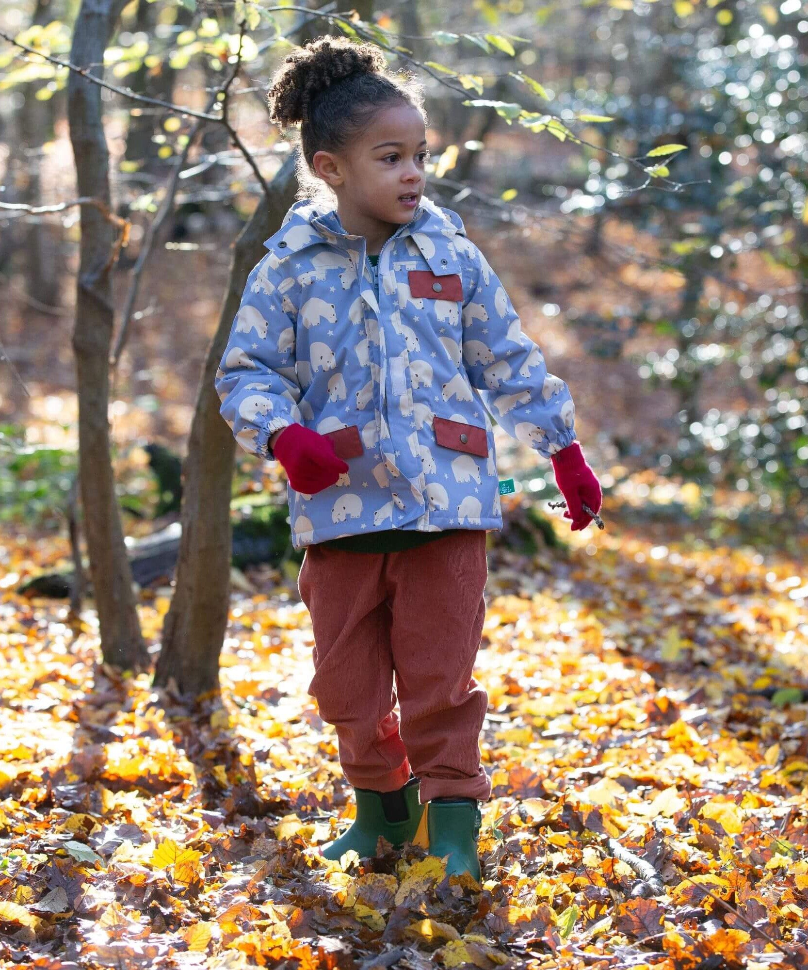 A child walking through leaves in a woodland playing with sticks and wearing the Little Green Radicals comfy corduroy trousers in ginger.  A corduroy loose fitted trousers in dark orange organic cotton fabric