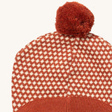 A close up of the Little Green Radicals ginger diamond knitted beanie hat showing the diamond design detail and dark orange pom pom on organic cotton fabric.