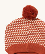 A close up of the Little Green Radicals ginger diamond knitted beanie hat showing the diamond design detail and dark orange pom pom on organic cotton fabric.