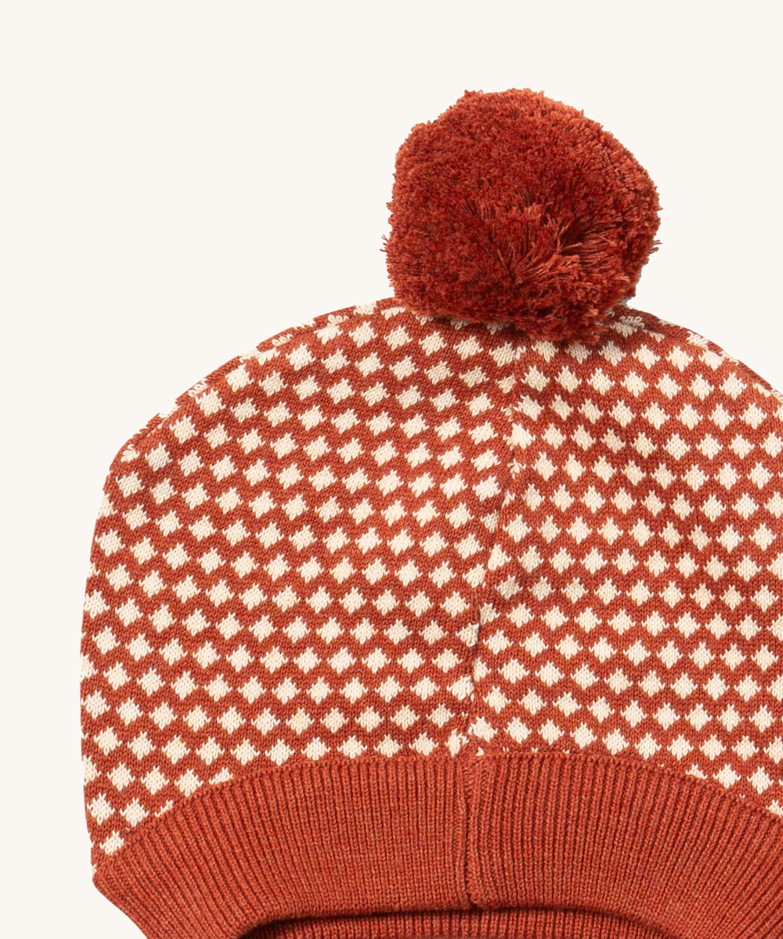 A close up of the Little Green Radicals ginger diamond knitted beanie hat showing the diamond design detail and dark orange pom pom on organic cotton fabric.