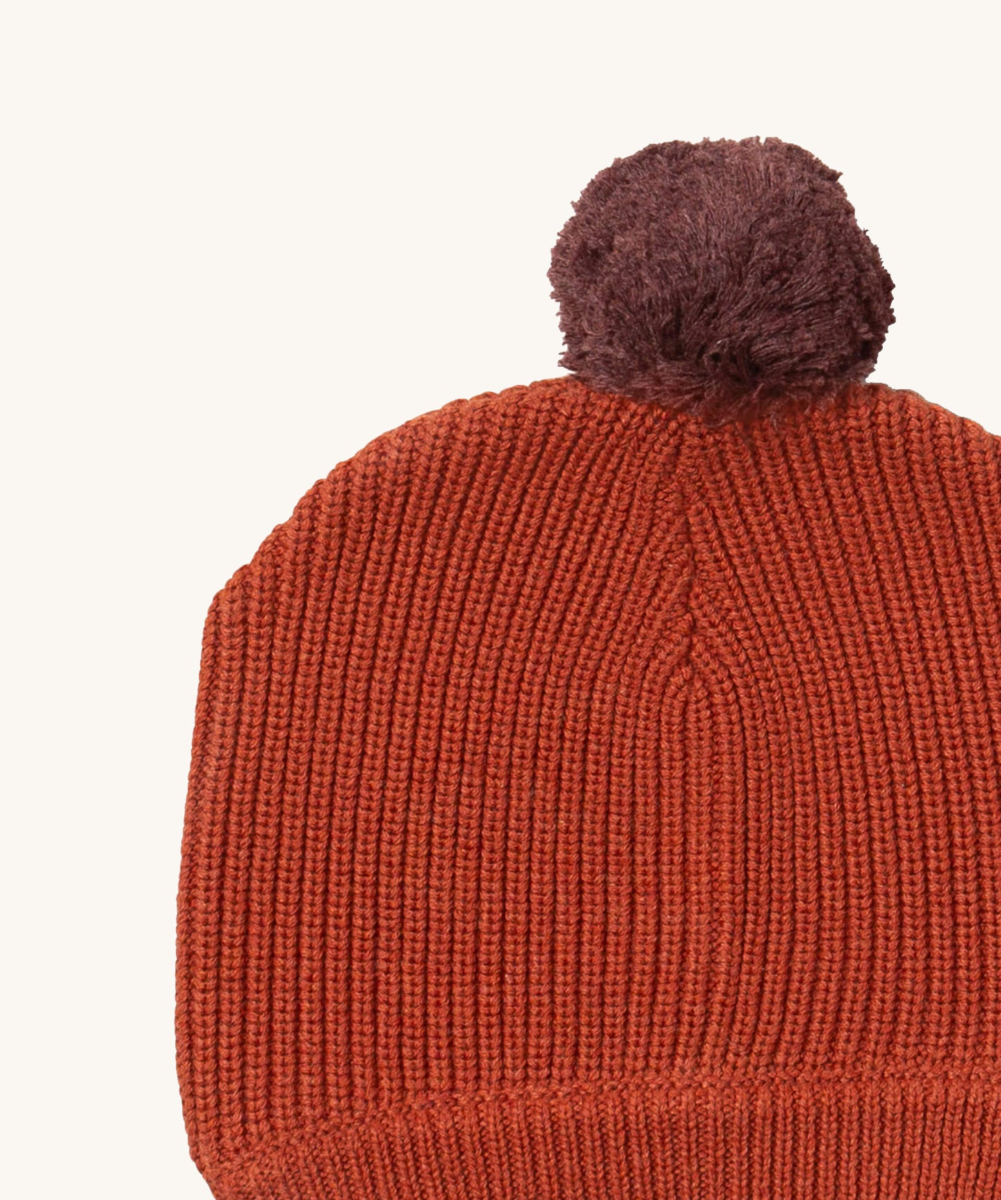 A close up of the Little Green Radicals ginger knitted beanie hat showing the knitted detail and purple pom pom on dark orange organic cotton fabric.