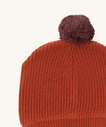 A close up of the Little Green Radicals ginger knitted beanie hat showing the knitted detail and purple pom pom on dark orange organic cotton fabric.