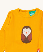 A close up of the Little Green Radicals gold brown owl applique long sleeve top showing the applique and button fastener detail. Features a small mushroom applique on the sleeve on dark yellow organic cotton fabric