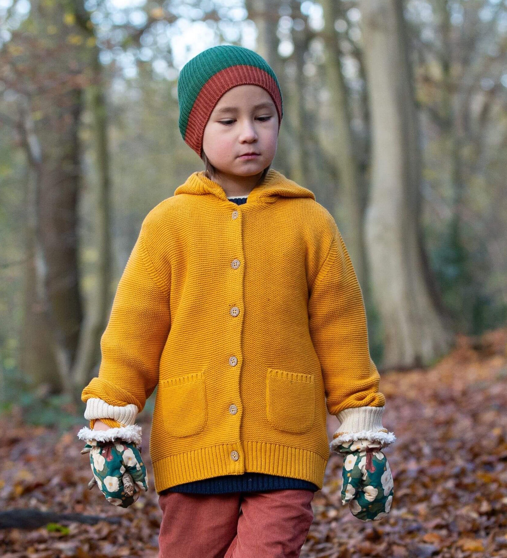 A child standing in woodland wearing the Little Green Radicals gold snuggly hooded knitted cardigan available at Babipur. A thick knitted cardigan with 2 pouch side pockets and button fasteners on yellow organic cotton fabric. 