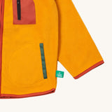 A close up of the Little Green Radicals polar fleece jacket in gold showing the chest and pocket detail and a close up of the sun appliqué patch on the shoulder. 