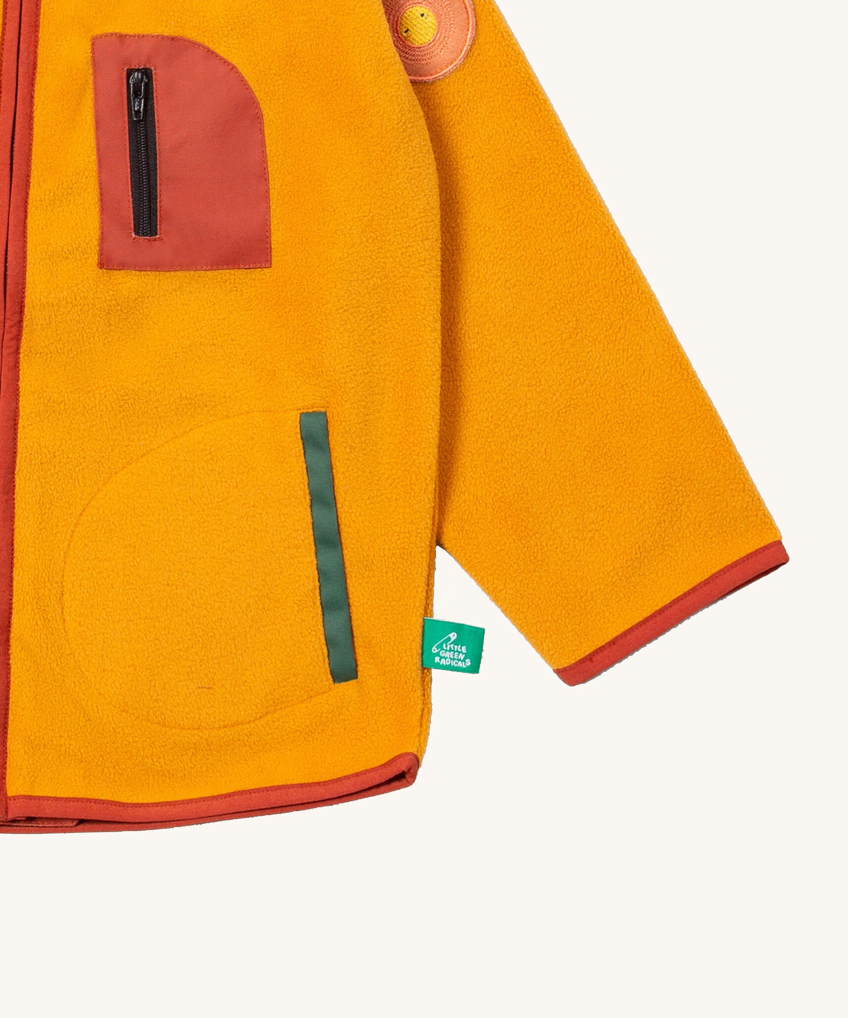 A close up of the Little Green Radicals polar fleece jacket in gold showing the chest and pocket detail and a close up of the sun appliqué patch on the shoulder. 