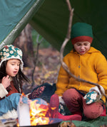 A child outside in woodland wearing the Little Green Radicals gold snuggly hooded knitted cardigan. A knitted cardigan with a generous hood for warmth and button fasteners down the middle on yellow organic cotton fabric. 