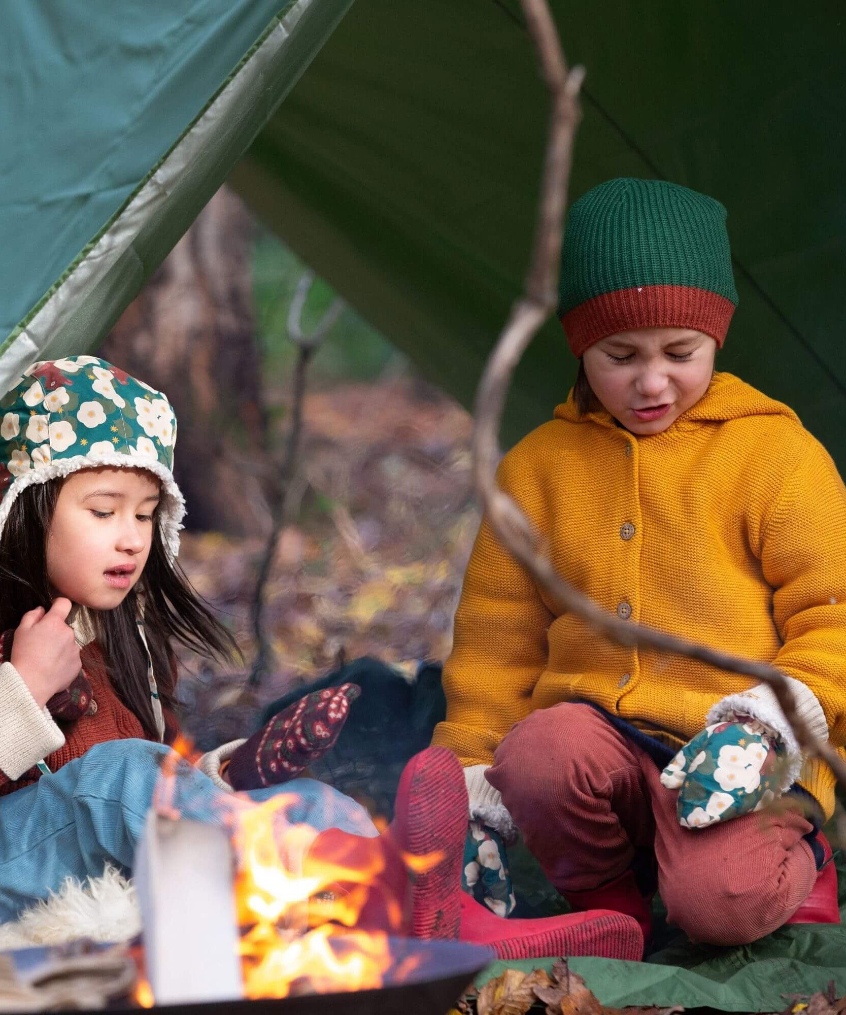 A child outside in woodland wearing the Little Green Radicals gold snuggly hooded knitted cardigan. A knitted cardigan with a generous hood for warmth and button fasteners down the middle on yellow organic cotton fabric. 