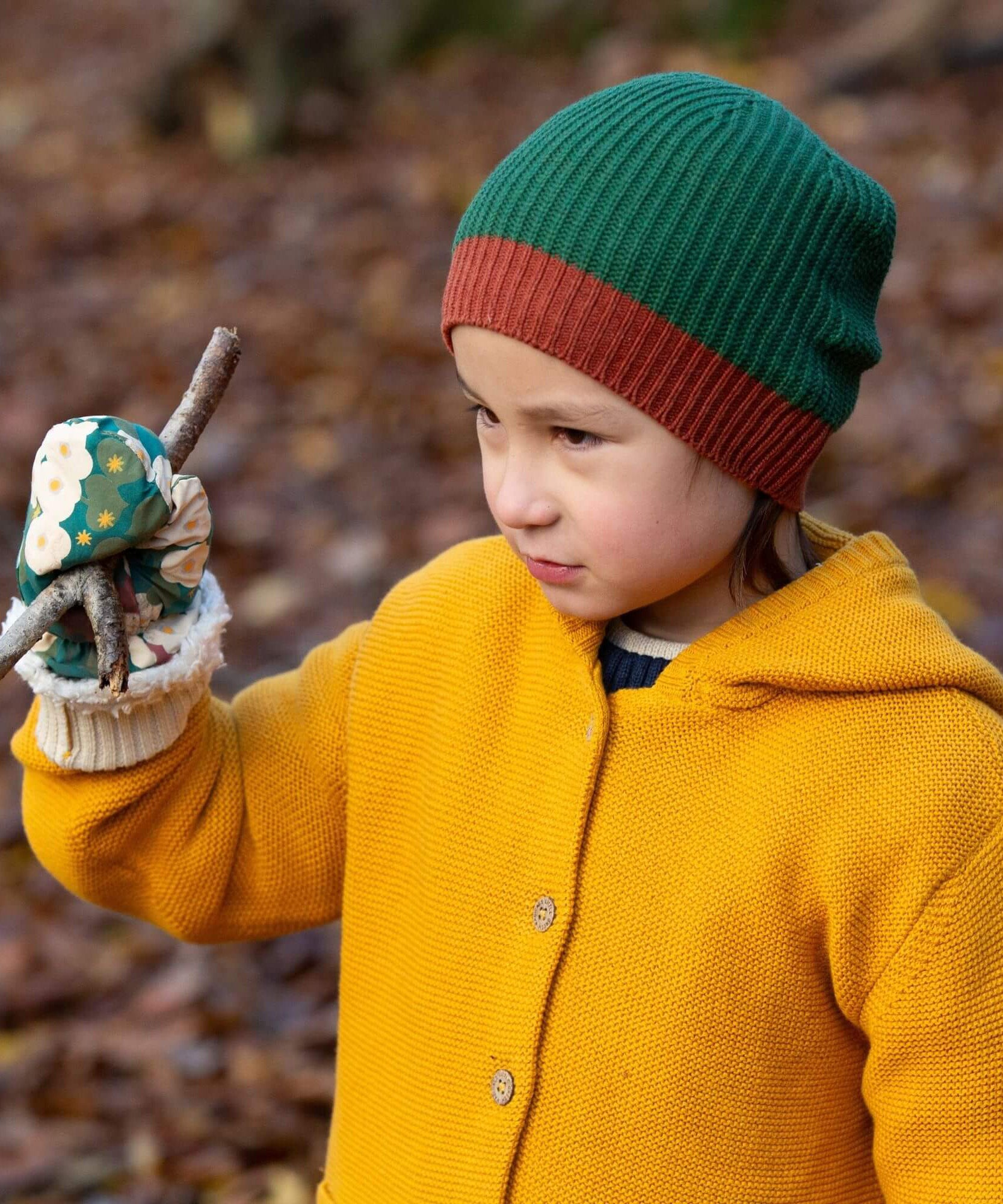 A child in woodland wearing the Little Green Radicals gold snuggly hooded knitted cardigan. A thick knitted cardigan with a generous hood for warmth and button fasteners down the middle with a cream cuff on yellow organic cotton fabric. 