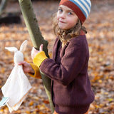A child standing next to a tree and wearing the Little Green Radicals knitted cardigan in chocolate. A warm brown GOTS organic cotton cardigan with yellow arm cuffs