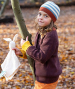 A child standing next to a tree and wearing the Little Green Radicals knitted cardigan in chocolate. A warm brown GOTS organic cotton cardigan with yellow arm cuffs