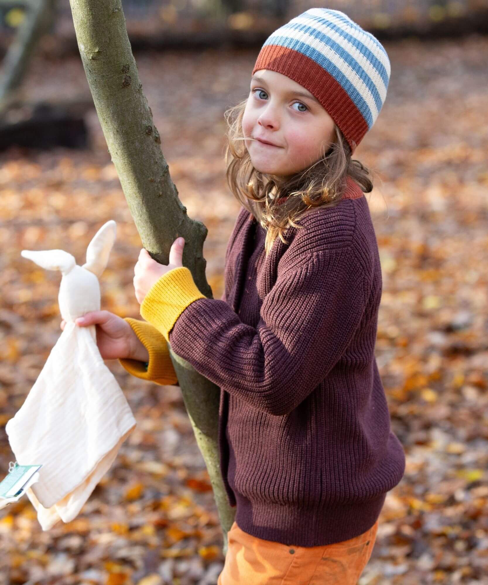A child standing next to a tree and wearing the Little Green Radicals knitted cardigan in chocolate. A warm brown GOTS organic cotton cardigan with yellow arm cuffs