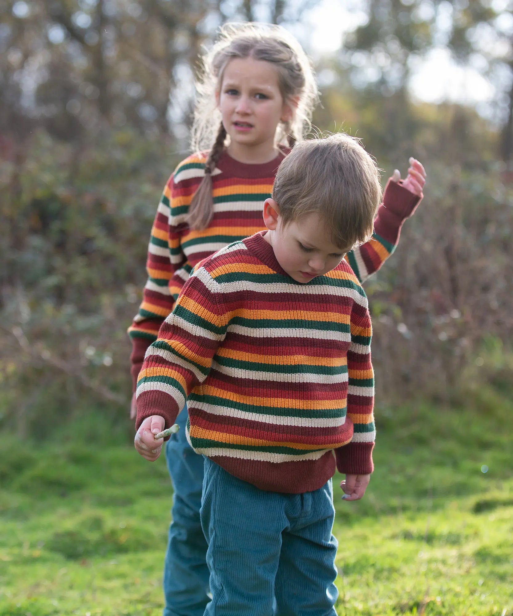Children wearing the Little Green Radicals knitted snuggly jumper in a  hazel rainbow striped design  with blue corduroy trousers. 
