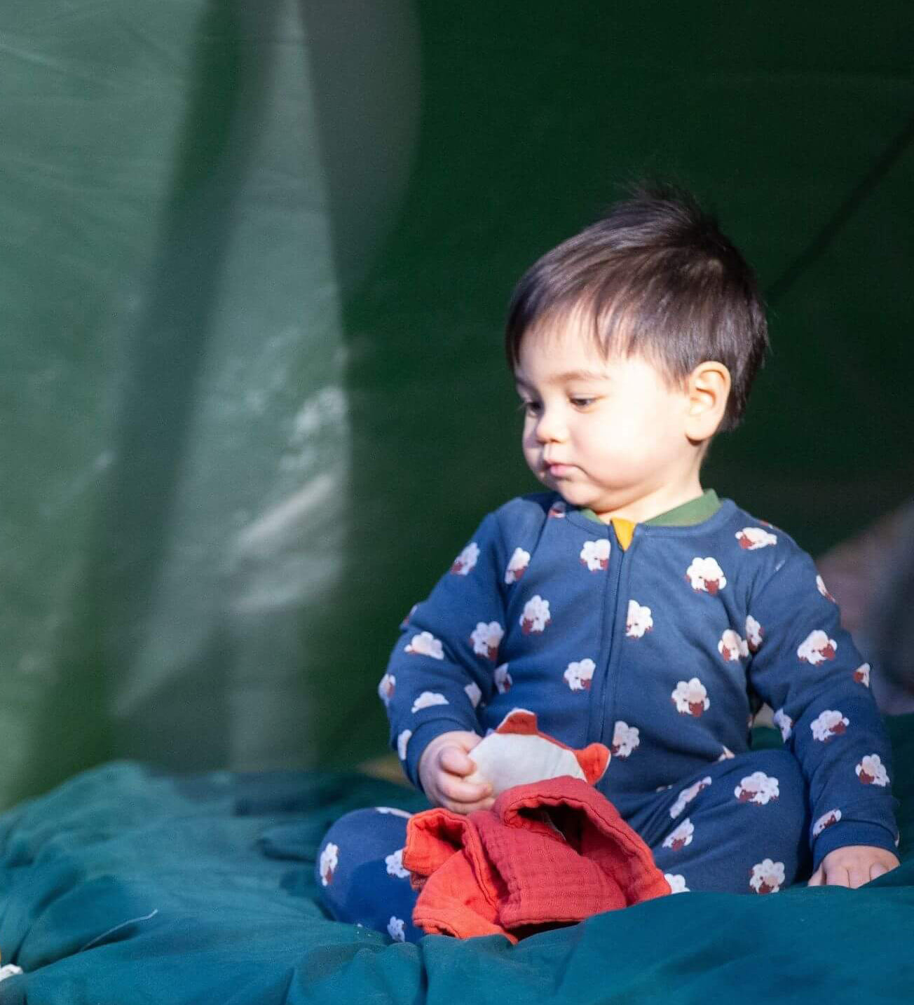 A child sitting under a shelter outside and wearing the Little Green radicals navy sheep baby grow. A soft GOTS organic cotton all in one in navy with cute sheep prints. Features a full length zip fastener with a yellow toggle 