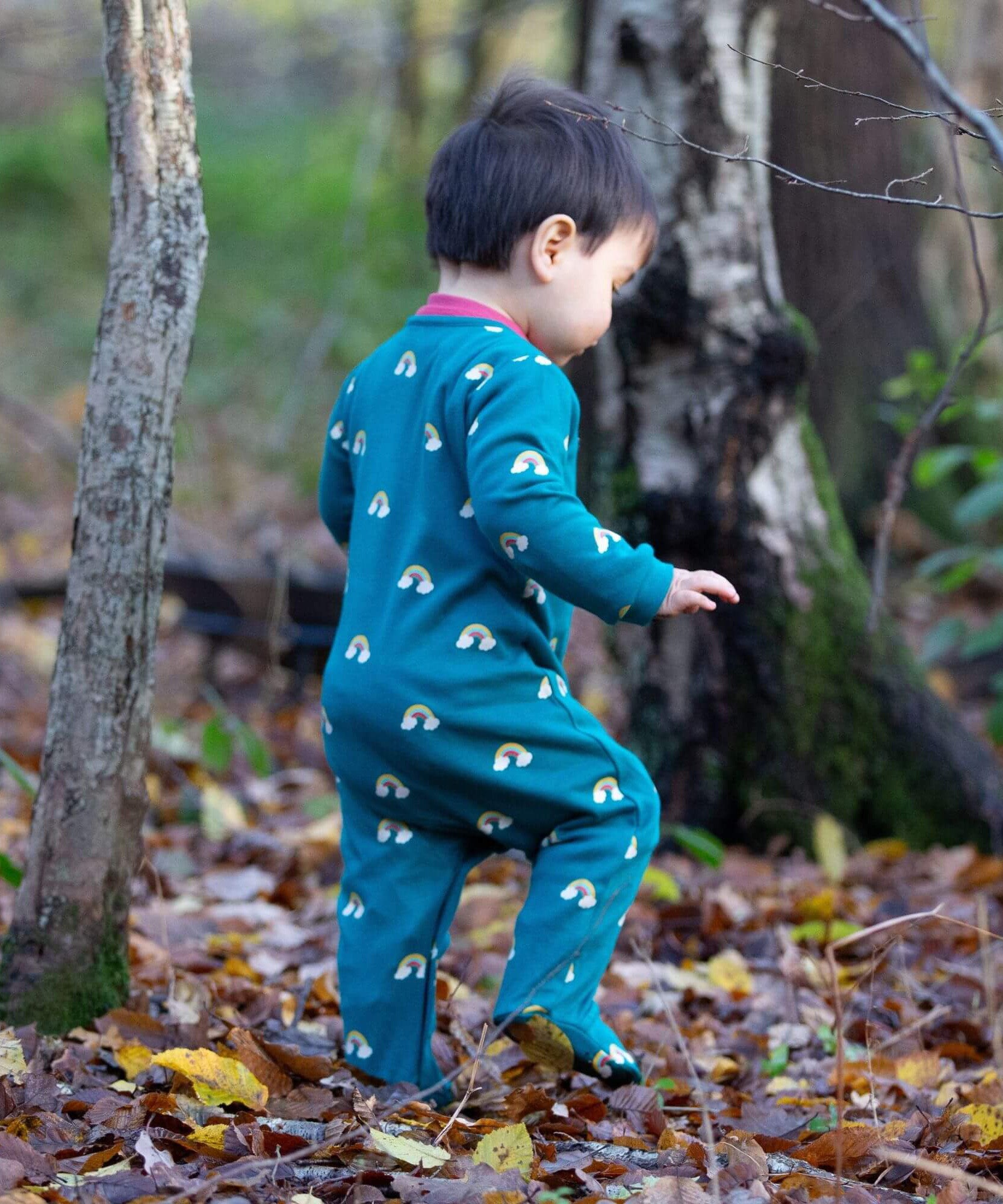 A child walking in the woods and wearing the Little Green radicals turquoise rainbow baby grow. A soft GOTS organic cotton all in one in turquoise with cute rainbow prints