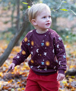 A child playing in woodland and wearing the Little Green Radicals flying birds quilted sweatshirt that's available at Babipur. A thick quilted sweatshirt with cute prints of birds flying through a starry night on dark maroon organic cotton fabric.
