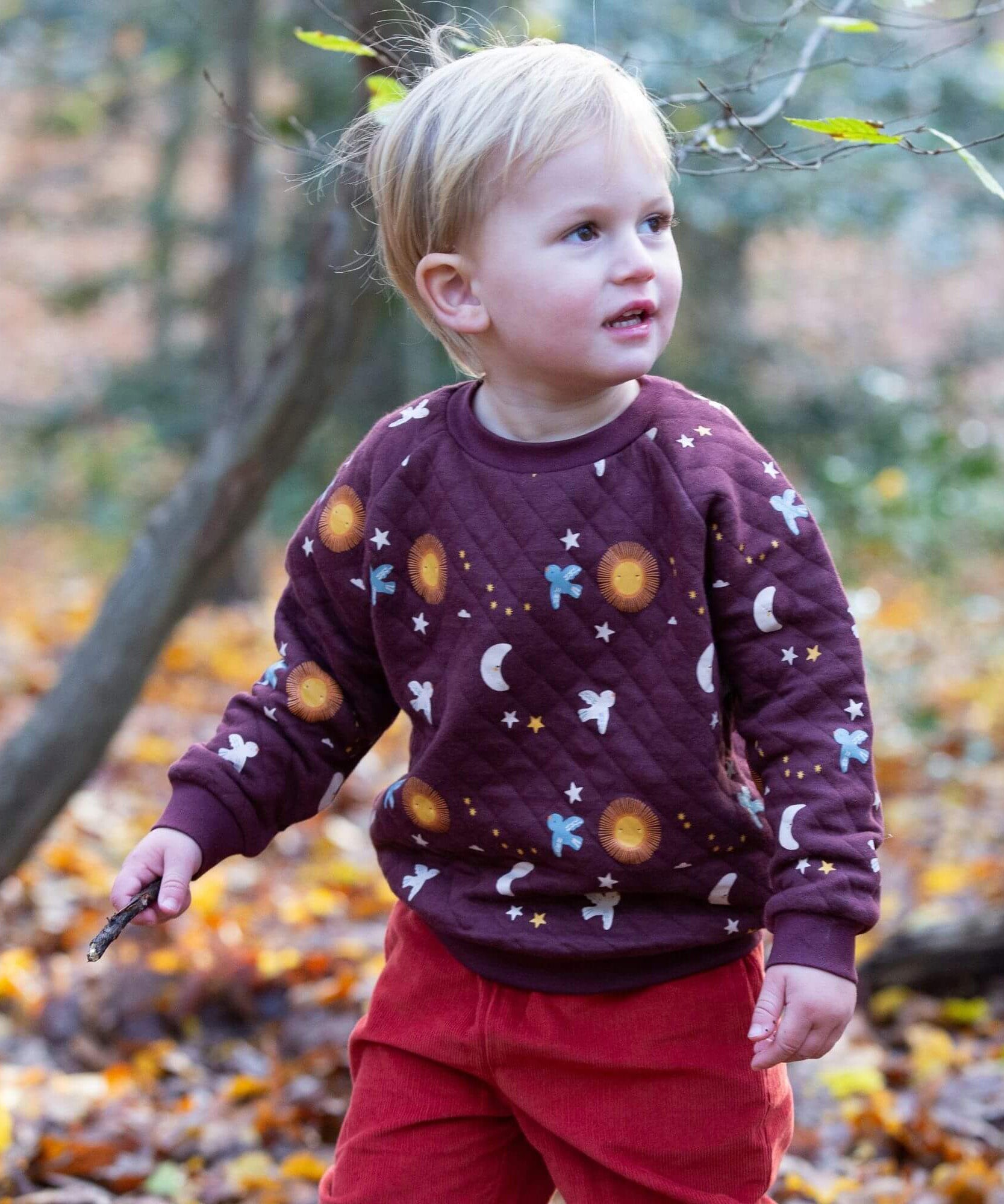 A child playing in woodland and wearing the Little Green Radicals flying birds quilted sweatshirt that's available at Babipur. A thick quilted sweatshirt with cute prints of birds flying through a starry night on dark maroon organic cotton fabric.