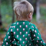 A child standing in woodland and wearing  the Little Green Radicals brown owl raglan sweatshirt that's available at Babipur. A soft knit long sleeve sweatshirt with cute brown on green organic cotton fabric.