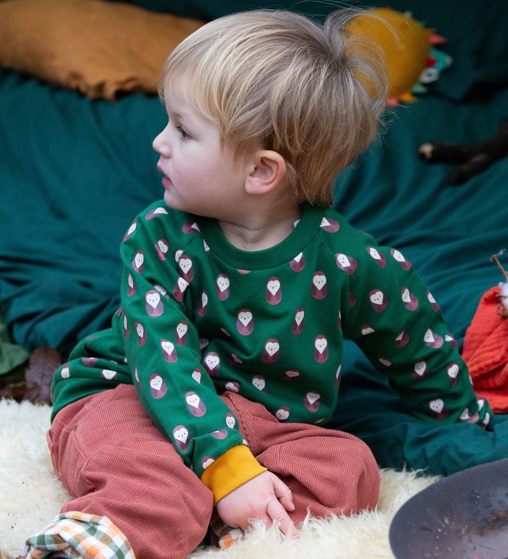 A child sitting outside and wearing the Little Green Radicals brown owl raglan sweatshirt that's available at Babipur. A soft knit long sleeve sweatshirt with cute brown owl prints and yellow trim on green organic cotton fabric.