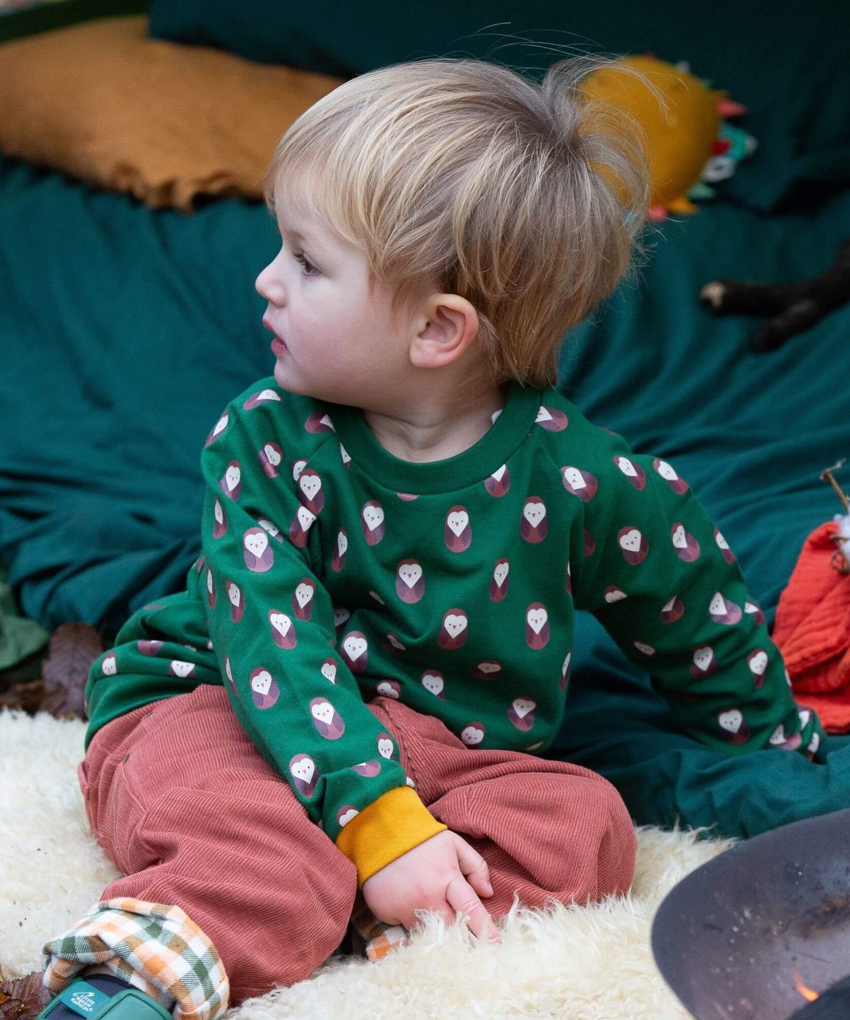 A child sitting outside and wearing the Little Green Radicals brown owl raglan sweatshirt that's available at Babipur. A soft knit long sleeve sweatshirt with cute brown owl prints and yellow trim on green organic cotton fabric.