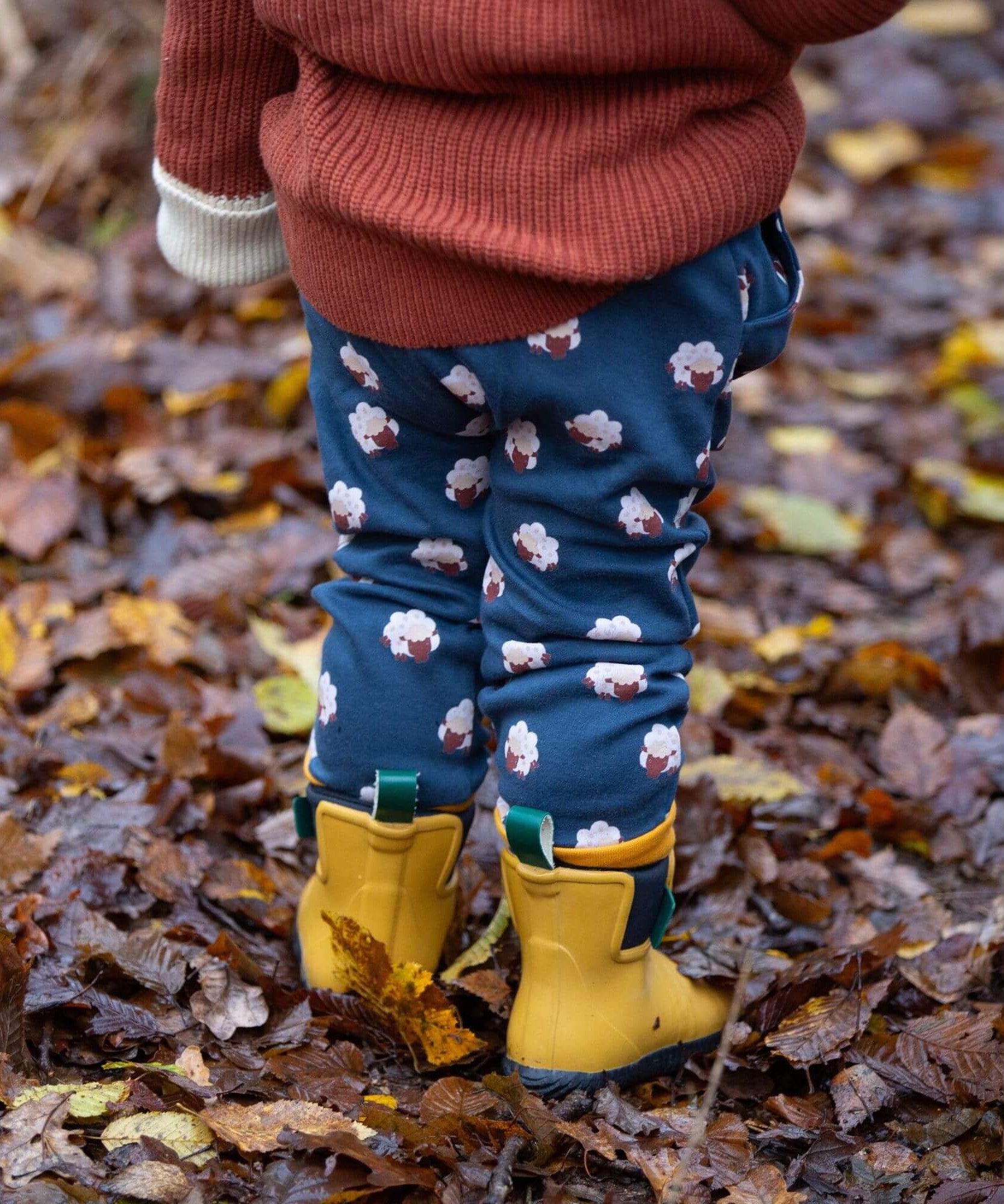 A child standing in  leaves in a woodland wearing the Little Green Radicals comfy joggers in sleeping sheep. A navy jogger with cute prints of sheep on soft GOTS organic cotton fabric