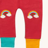 A close up of the Little Green Radicals soft red rainbow kneepatch joggers showing the rainbow embroidery kneepatch and the leg cuff detail on pale red organic cotton fabric.